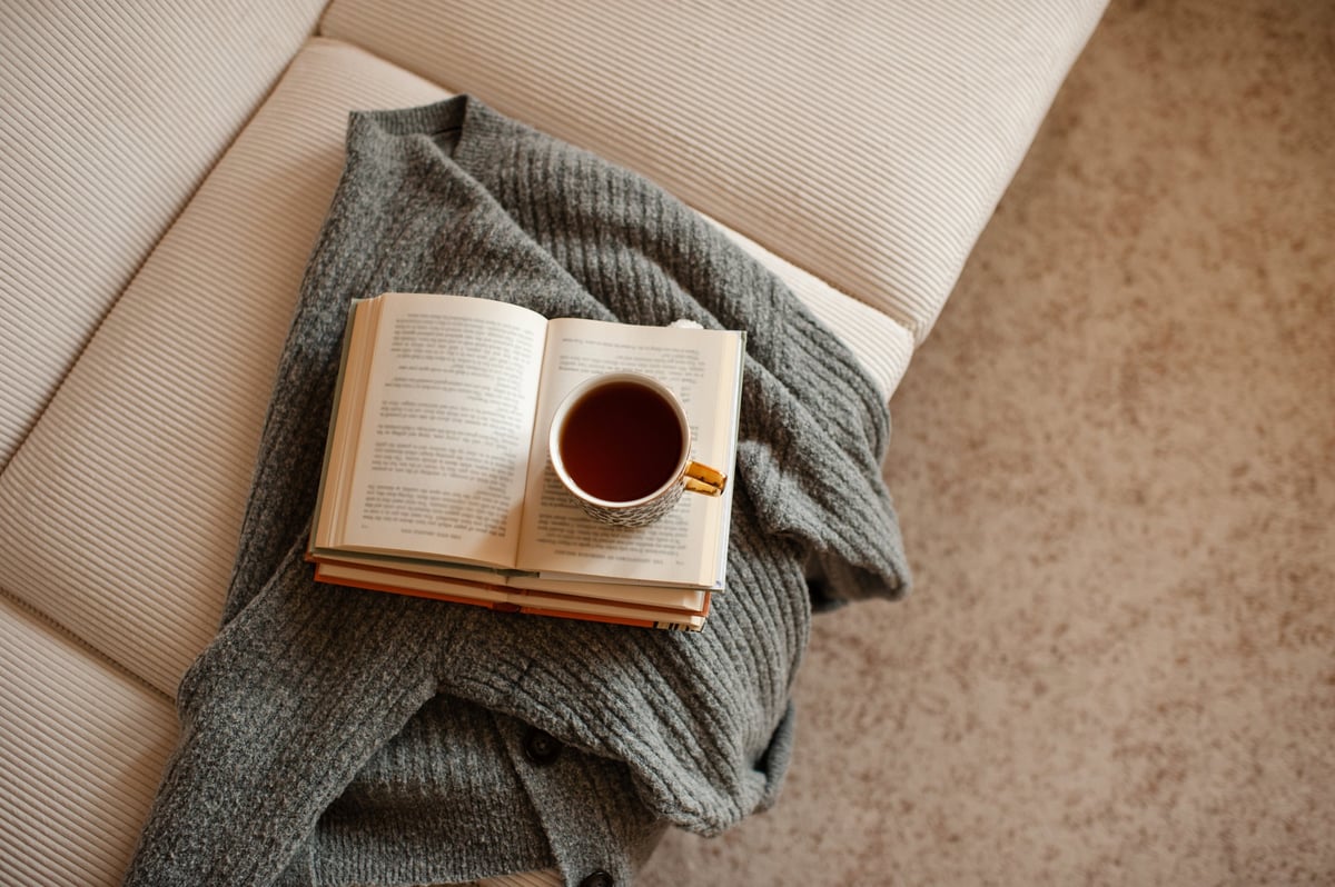 Cup of black tea staying on open paper books with knitted textile on white velvet couch in living room at home. Cozy and hygge atmosphere. Coziness.