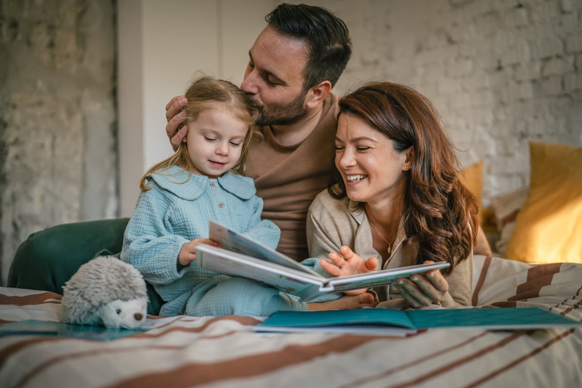 Happy parents and their young daughter are lying on a bed, bonding while reading a picture book and laughing aloud, sharing a joyful and intimate moment at home